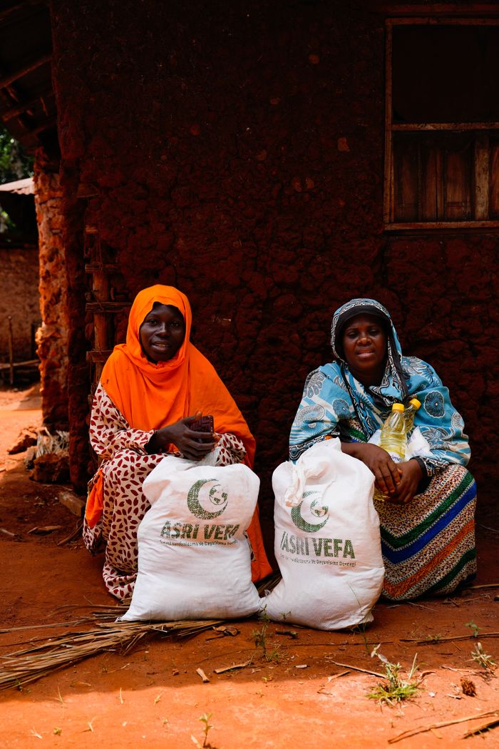 Two women sit by a mud house with relief bags, showing community aid.